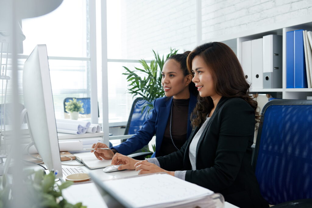 Two businesswomen sitting together in office at desk and looking at computer screen