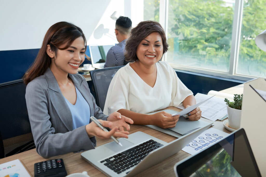 Two female colleagues sitting at desk in office with laptop helping each other