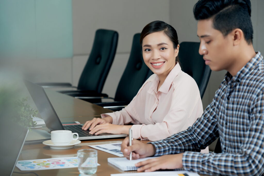 Woman sitting at table with laptop and male colleague writing in notebook