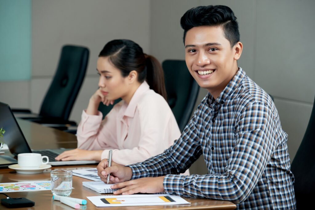 Young man sitting at meeting table in office and smiling, and woman working on laptop