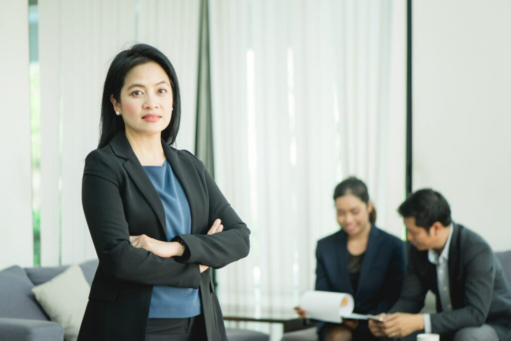 A confident manager standing with crossed arms while two colleagues discuss documents in the background.