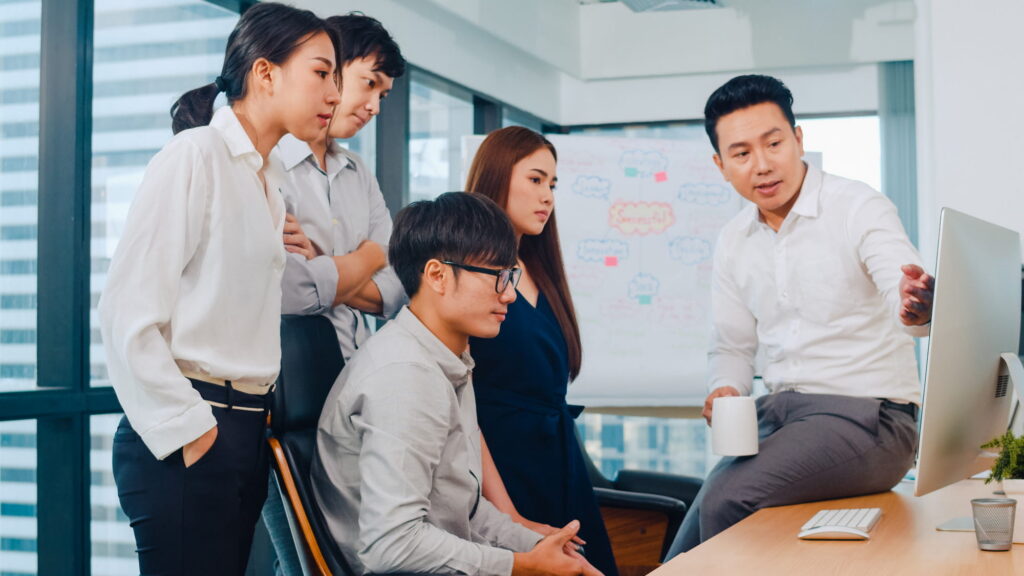 A corporate team gathers around an office desk while a man sitting on the edge explains the content on the computer.