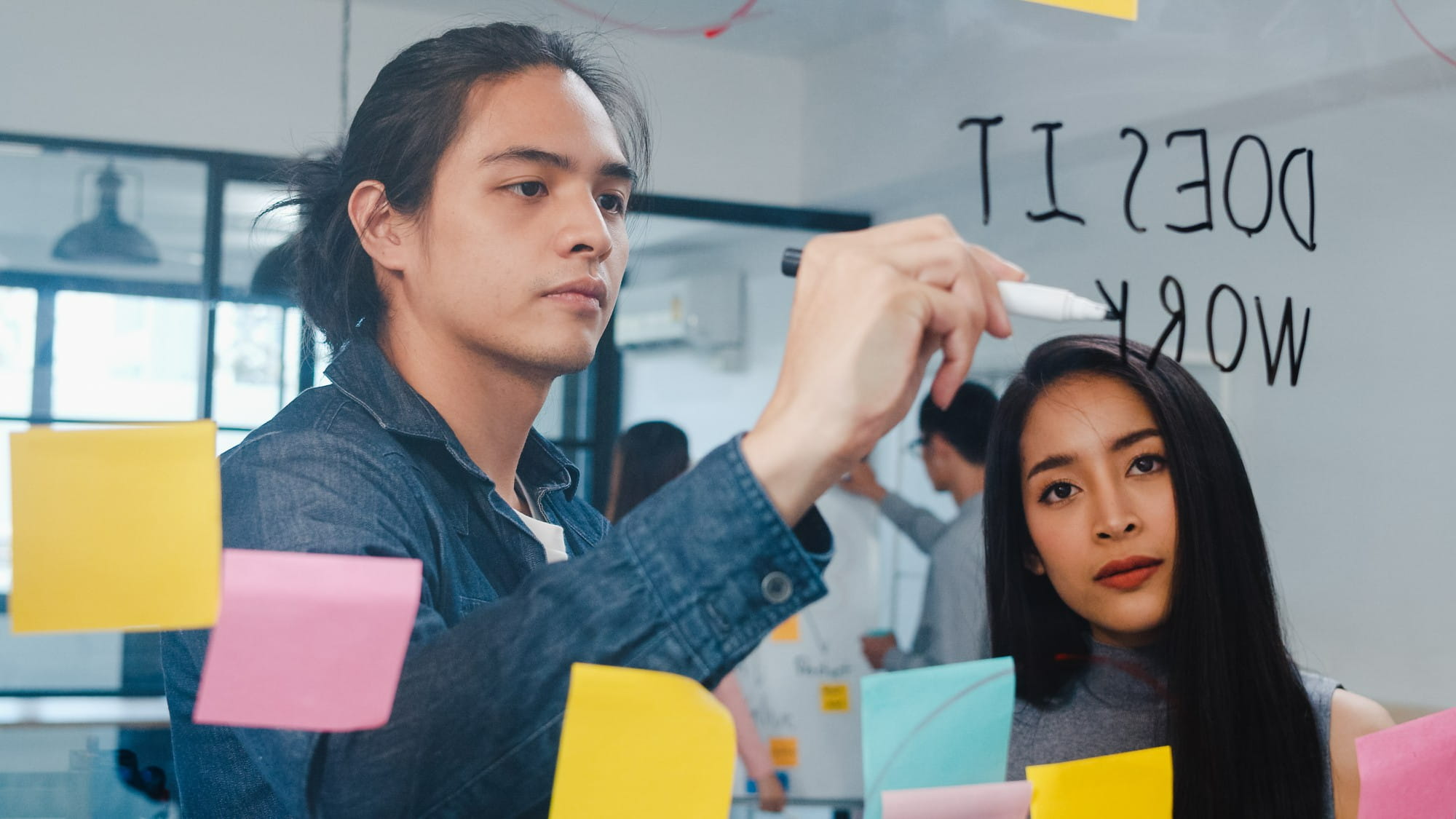 A man writing on a clear glass board with colorful sticky notes attached beside a woman in a collaborative office setting.