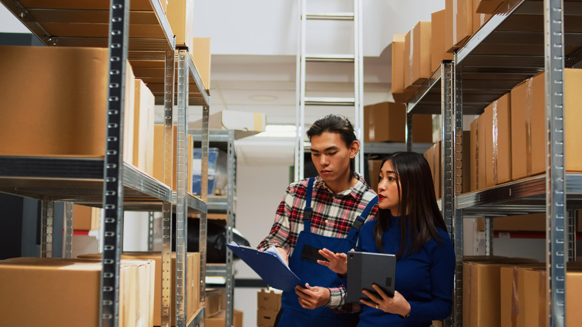 A manager and her staff checking the quality of inventory using a clipboard and a digital tablet between rows of warehouse shelves.