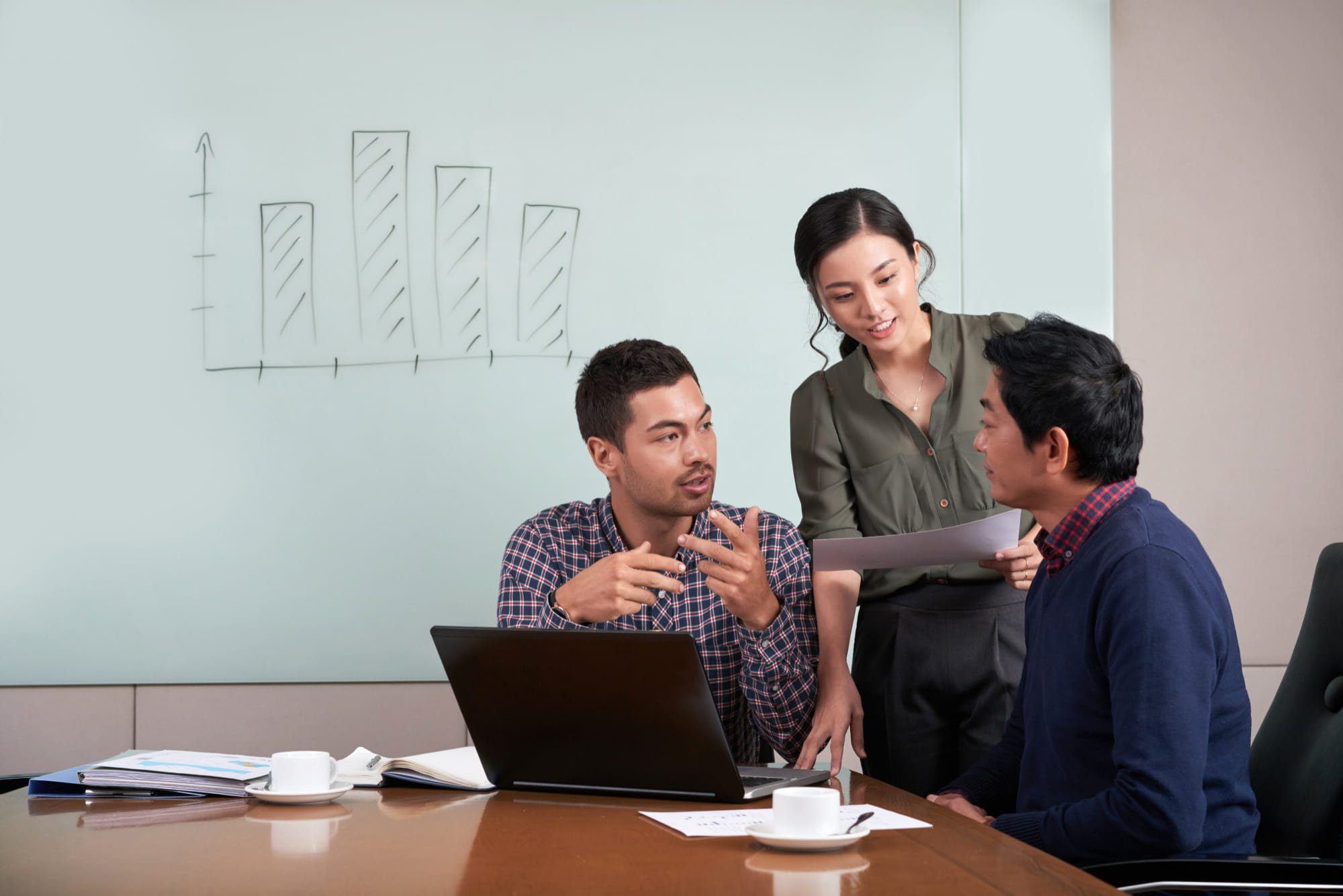 Three colleagues collaborate in a conference room around a laptop while a hand-drawn bar chart is visible on the whiteboard behind them.